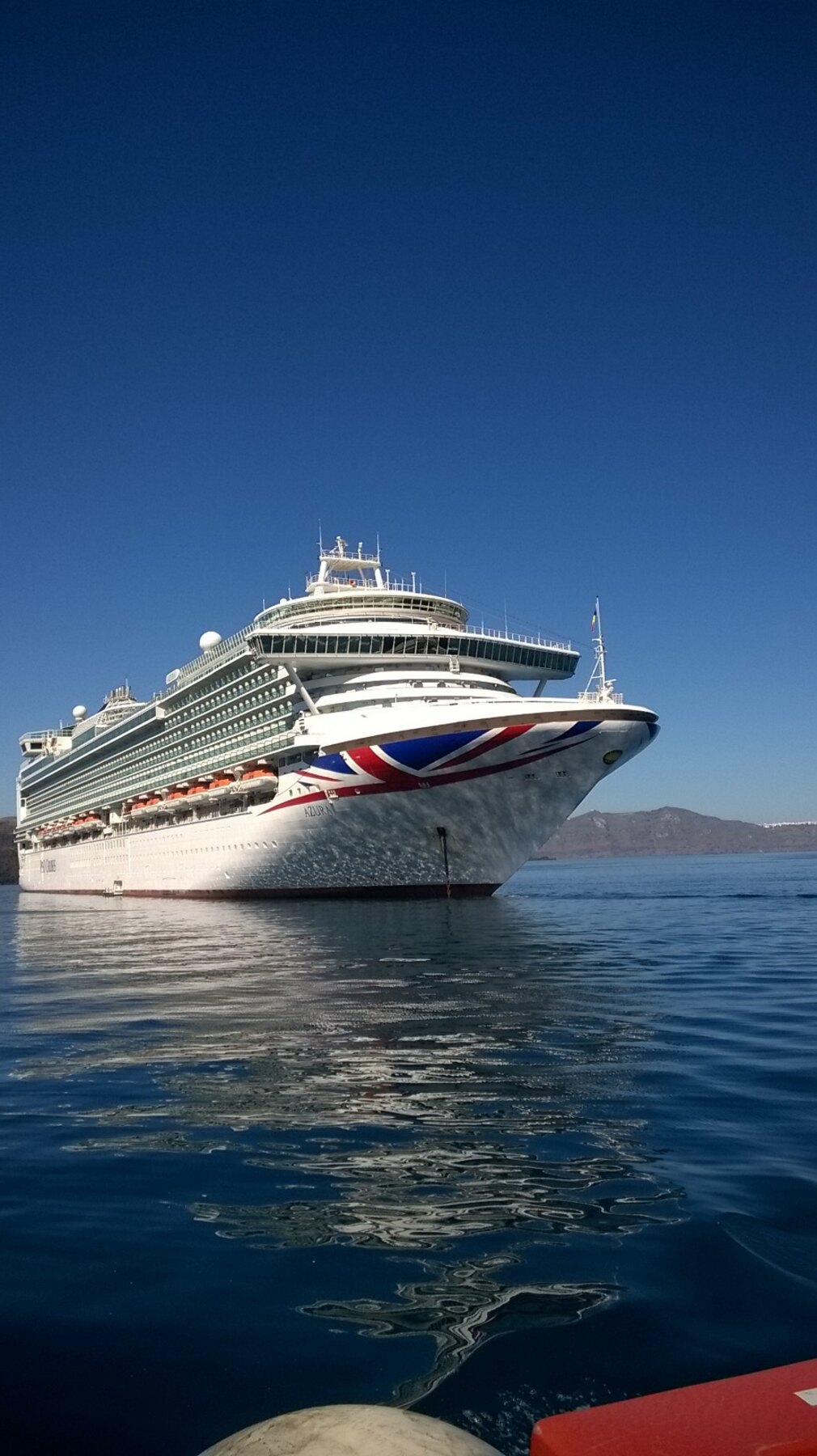 P&O's Azura at anchor in flat blue water, her Union Jack bow livery catching the sun.